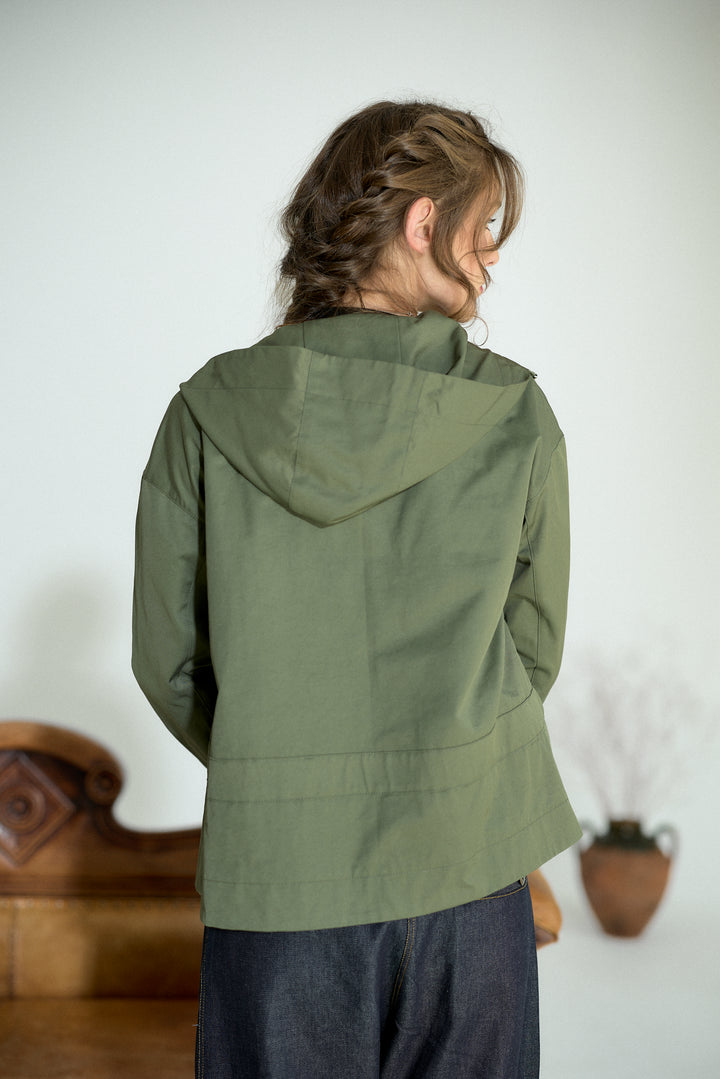 A woman viewed from behind wearing an olive green hooded jacket and dark blue jeans, standing in a minimalist indoor setting with a light gray wall and decorative wooden furniture in the background.