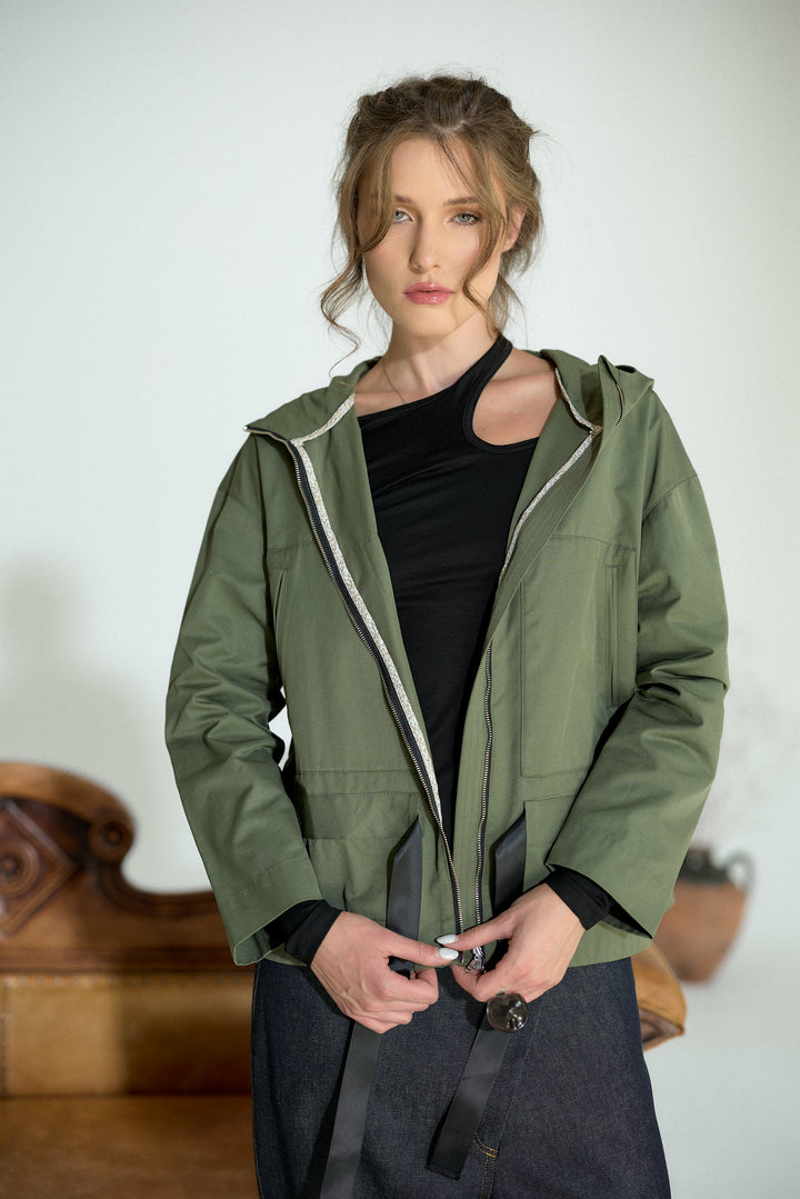 A woman wearing an olive green hooded jacket over a black top and dark skirt, standing indoors against a light wall.