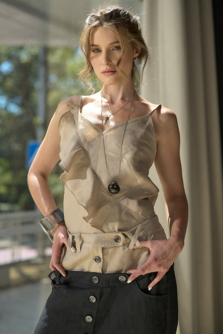 A woman wearing a beige ruffled sleeveless top and black pants, standing in a softly lit indoor setting with a window and curtains in the background.
