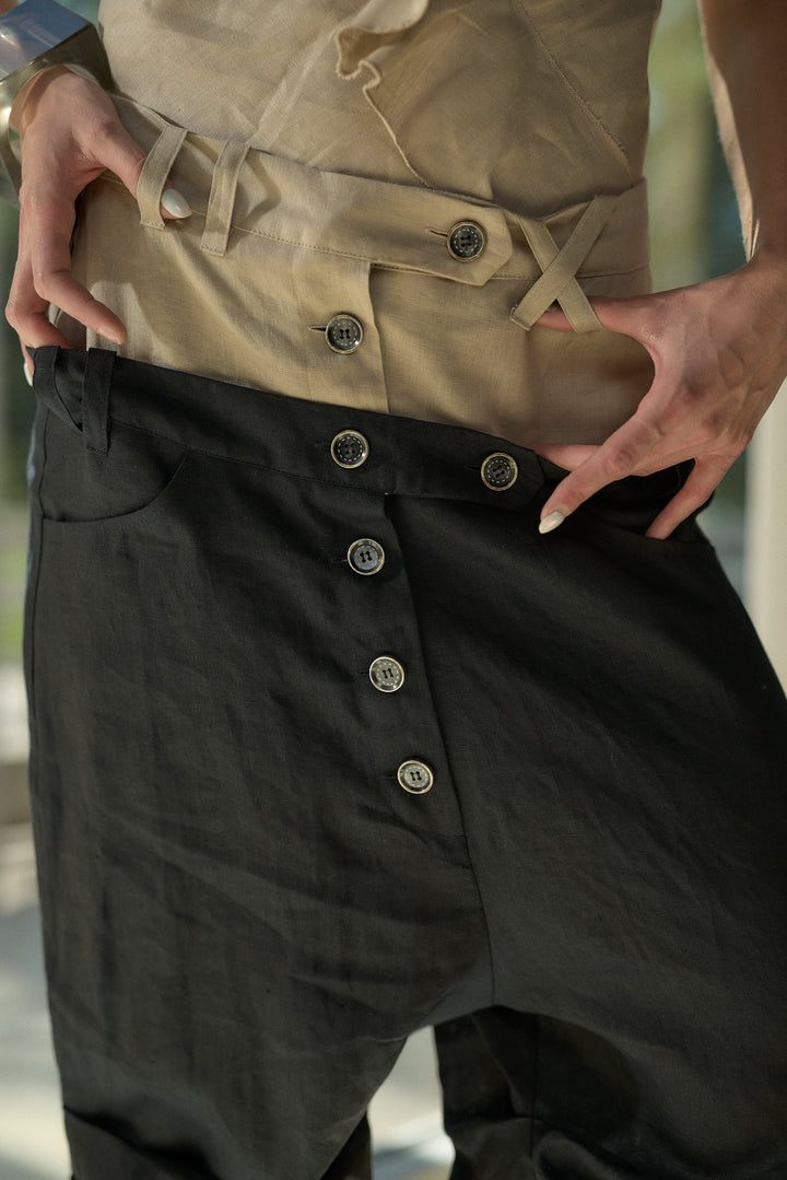 Close-up of a person wearing loose black pants with a vertical row of large silver buttons down the front, paired with a beige belted top. The person's hands are pulling the waistband of the pants slightly outward.