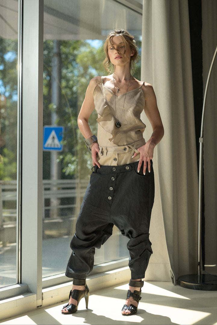A woman models a unique outfit consisting of a beige sleeveless top with a large central cutout and dark baggy pants, standing in a brightly lit room with large windows.