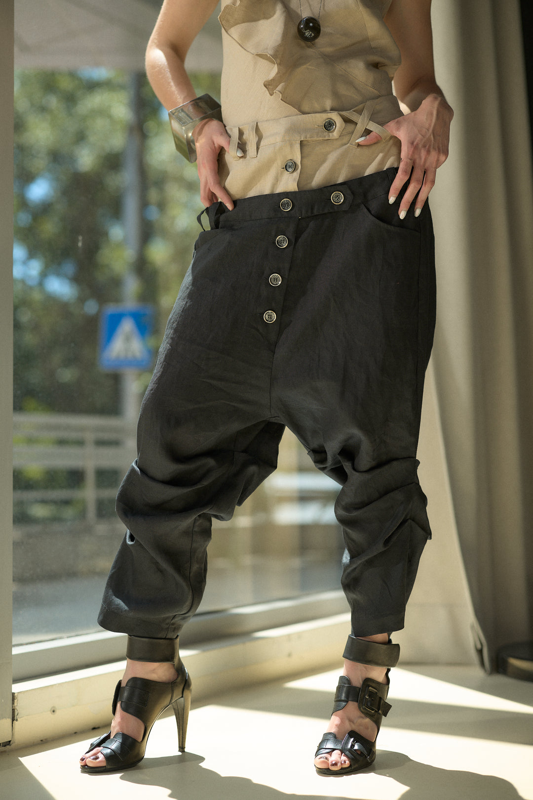 A person wearing loose black baggy pants with a high button fly, paired with a beige top and black high-heeled sandals, standing in front of a window.