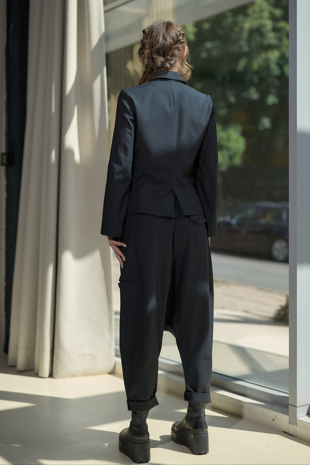 A person viewed from behind wearing a dark tailored suit with a structured blazer and loose cropped trousers, standing indoors near large windows and curtains.