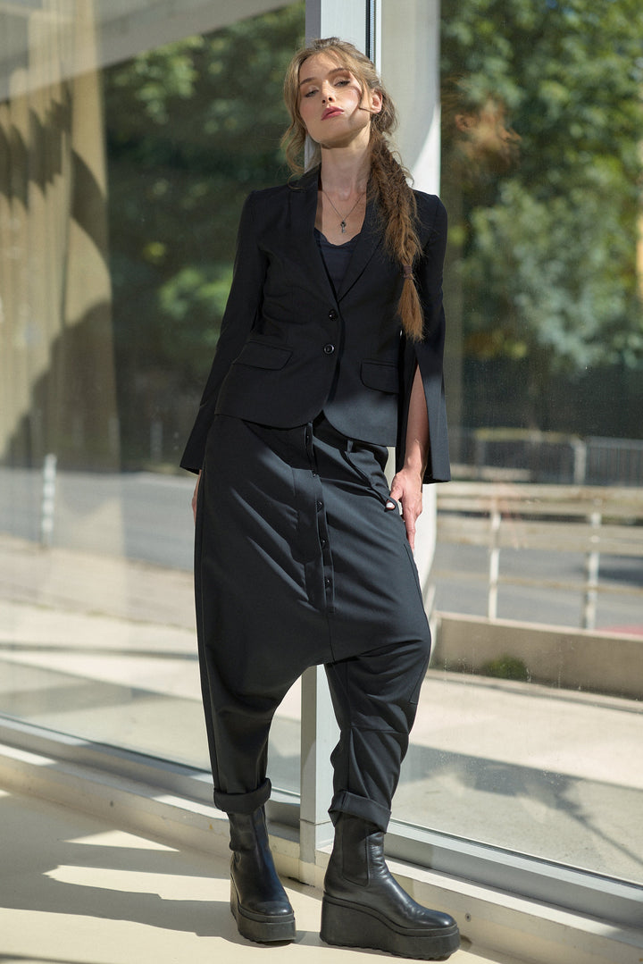 A woman stands by a glass storefront window wearing an all-black outfit consisting of a cropped blazer, long-sleeve top, baggy harem-style pants, and black platform boots.