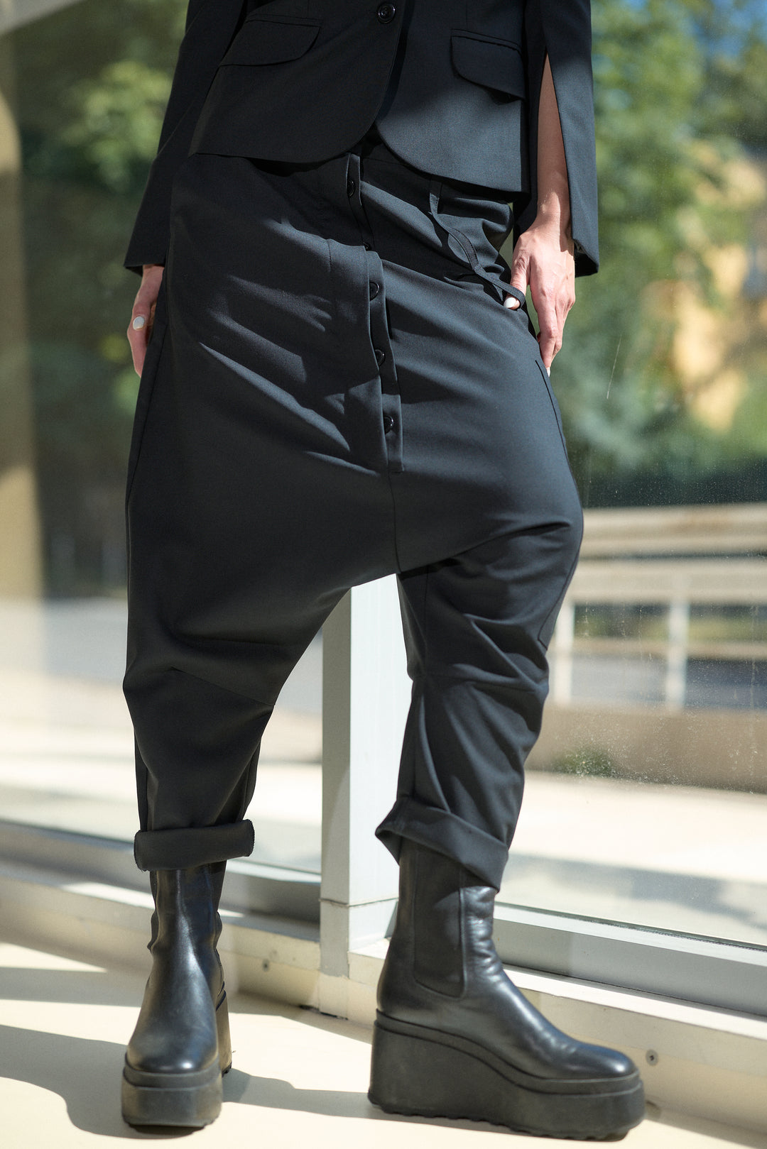 A person wearing loose black drape pants with a button front detail, paired with a black blazer and chunky black platform Chelsea boots, standing near a window.