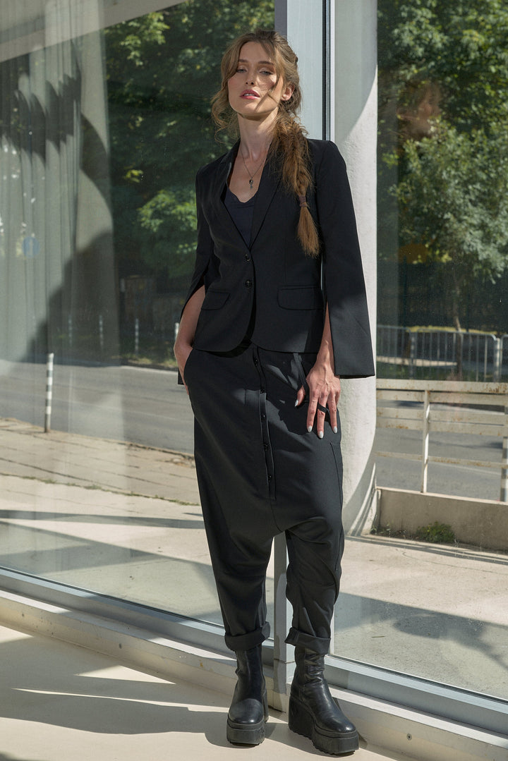 A woman stands outdoors beside a glass building, wearing a black tailored blazer with three-quarter sleeves, matching loose black trousers, and black ankle boots.