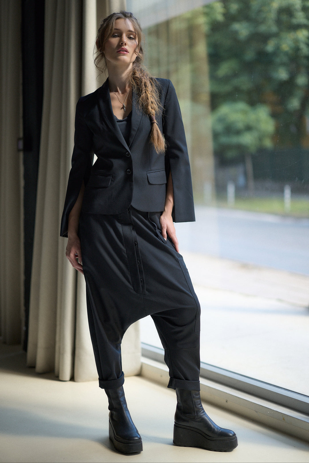 A woman models a black two-piece suit consisting of a cropped blazer with wide sleeves and matching relaxed-fit pants with a drawstring, paired with black platform ankle boots, standing by a large window.