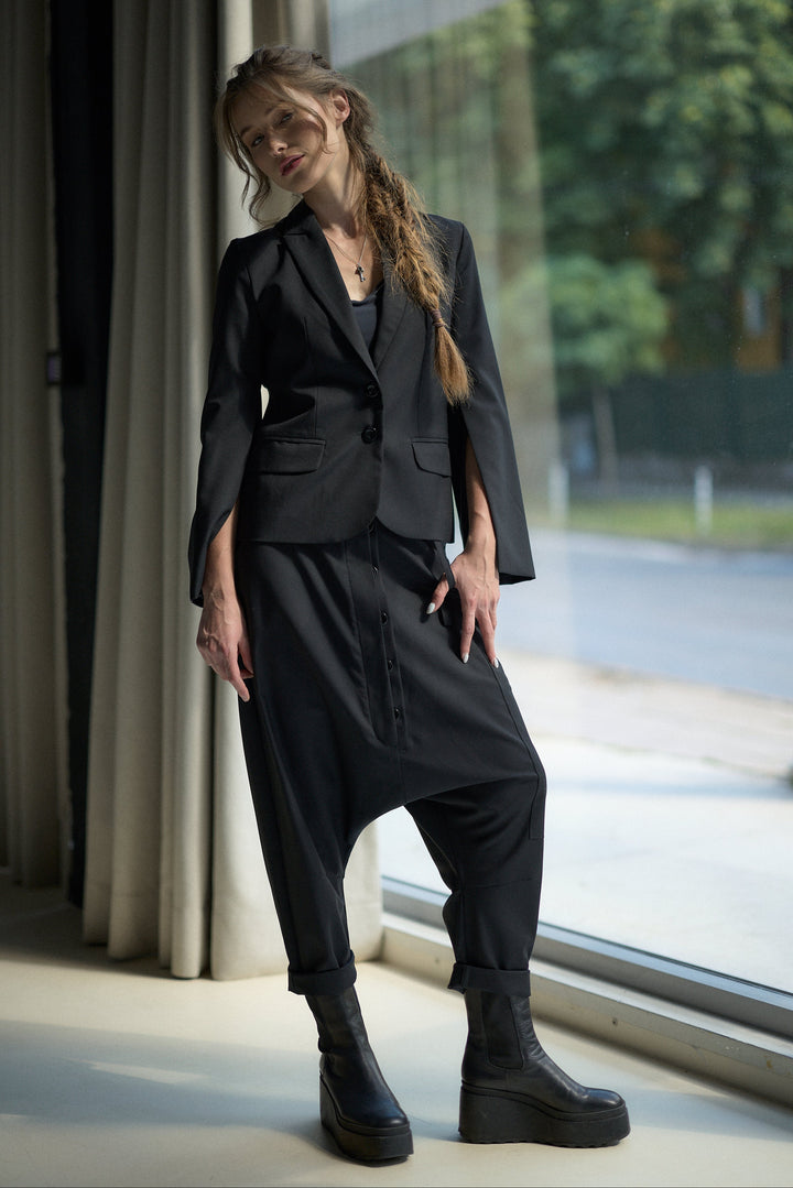 A woman models a black two-piece outfit consisting of a cropped blazer with wide cape-like sleeves and matching relaxed cargo-style pants, paired with black platform ankle boots, standing by a large window.
