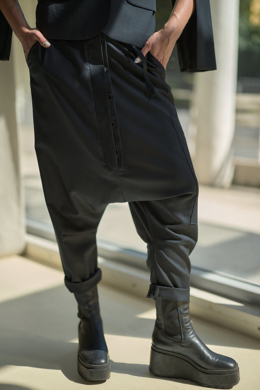 A person wearing loose black caged harem-style pants with a front button placket and cuffed ankles, paired with black platform ankle boots in a sunlit indoor setting.
