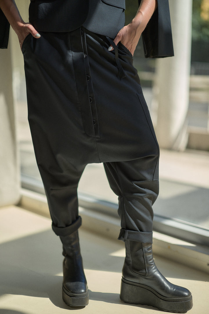 A person wearing loose black caged harem-style pants with a front button placket and cuffed ankles, paired with black platform ankle boots in a sunlit indoor setting.