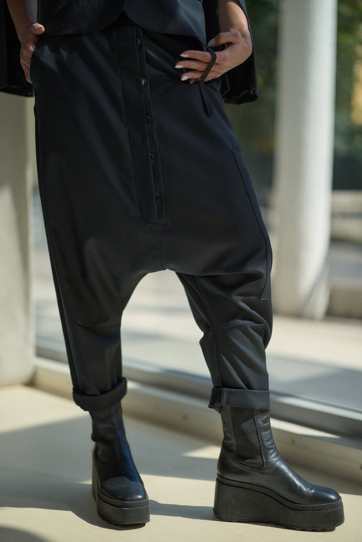 A person wearing loose black drape-style pants with a front button placket and black platform ankle boots, standing indoors near a window.