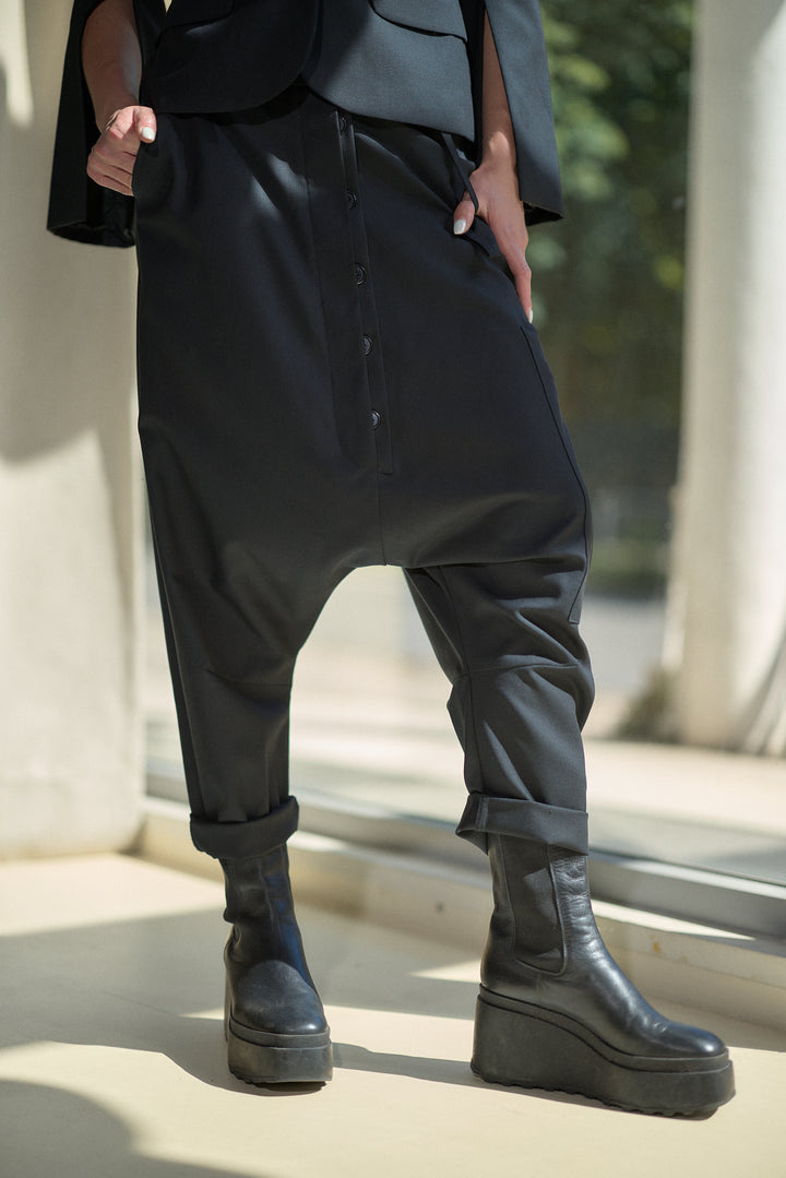 A person wearing loose black drape-style pants with cuffed hems, paired with black platform ankle boots, photographed in a sunlit architectural setting.
