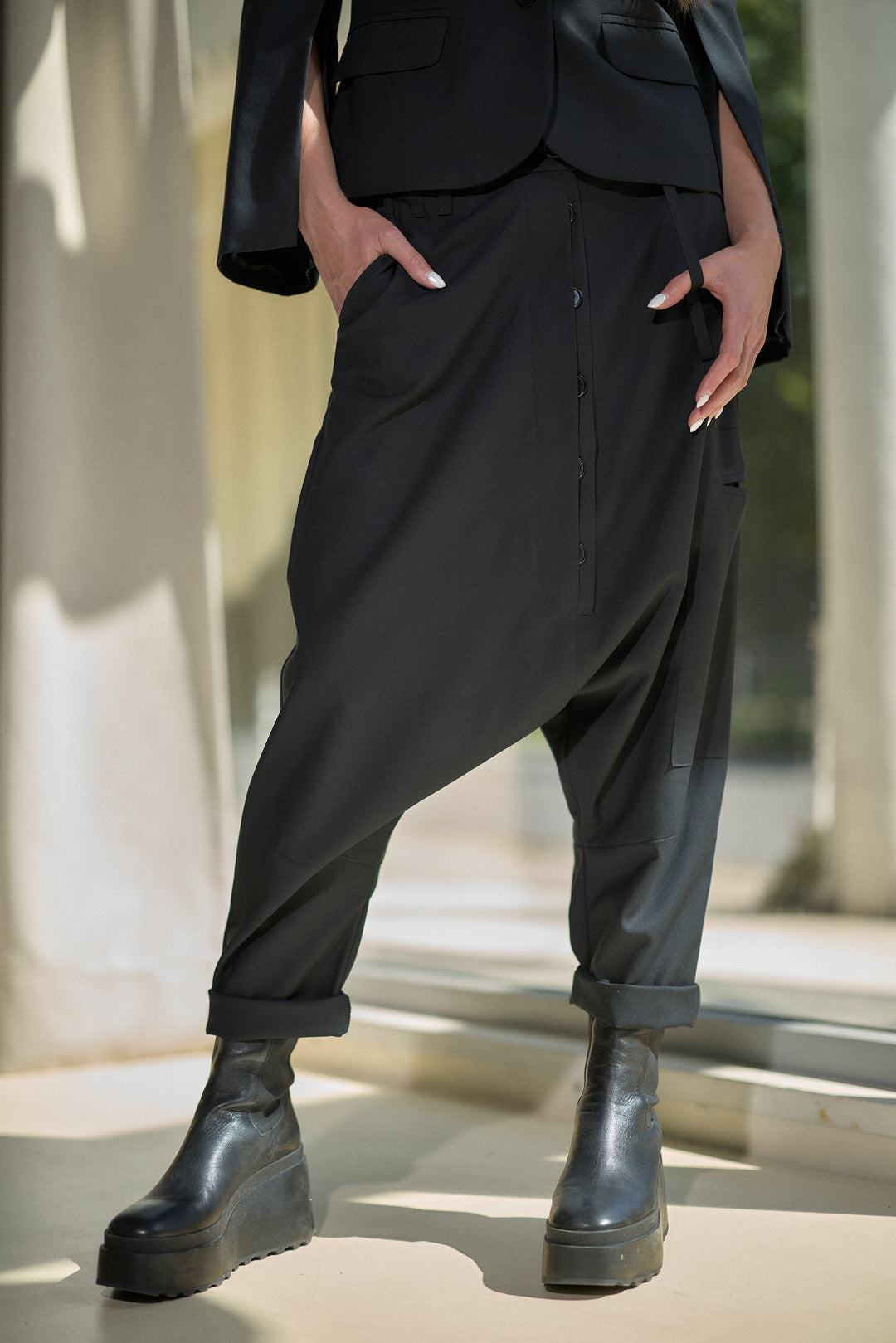 A person wearing loose black drape pants with a button front and cuffed hems, paired with black platform ankle boots, photographed in a sunlit architectural setting.