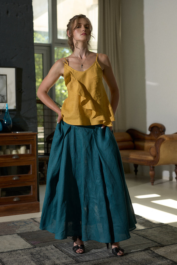 A woman stands in a sunlit room wearing a mustard yellow ruffled camisole top and a long teal skirt. The room has dark wood furniture, a large window, and a patterned rug.