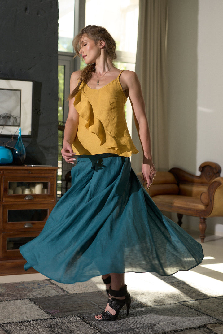 A woman modeling a two-piece outfit consisting of a mustard yellow ruffled spaghetti-strap top and a flowing teal blue skirt, standing in a sunlit room with a wooden cabinet and a sofa in the background.