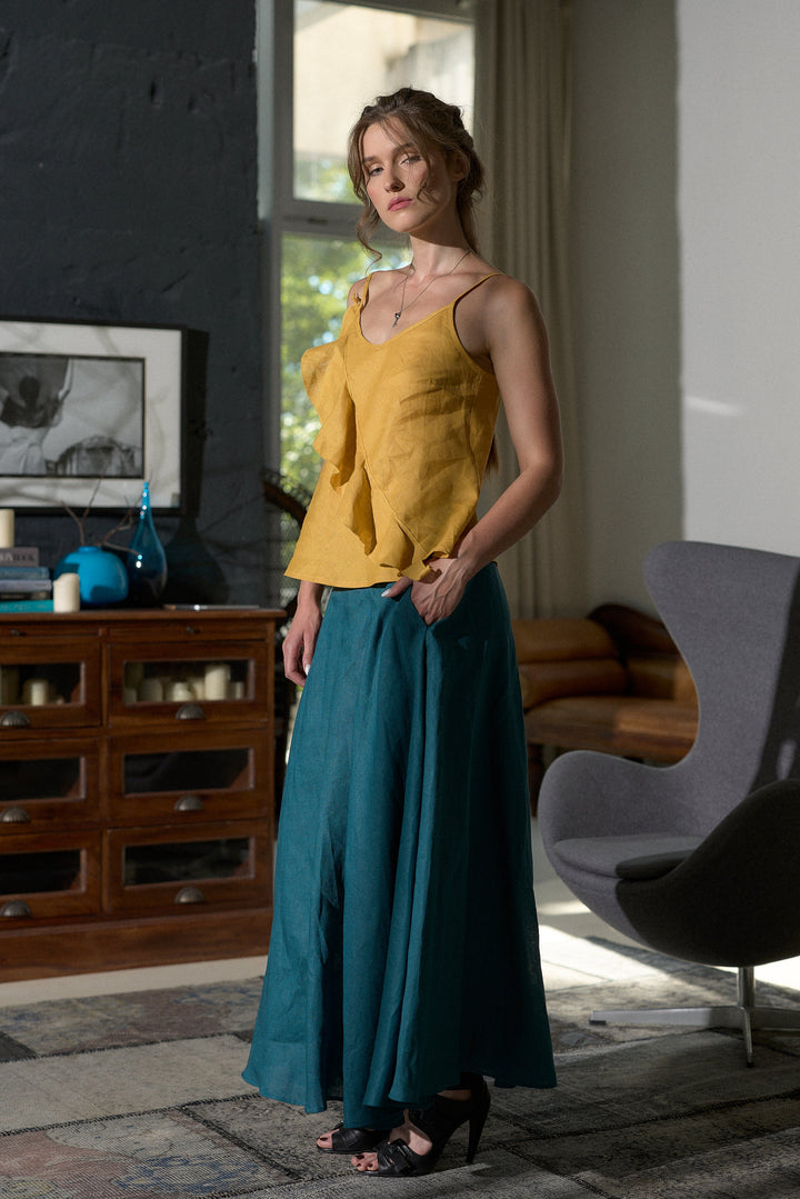 A woman modeling a two-piece outfit consisting of a mustard yellow ruffled tank top and a teal wide-leg skirt, standing in a modern room with a dark wood cabinet, gray chair, and textured walls.