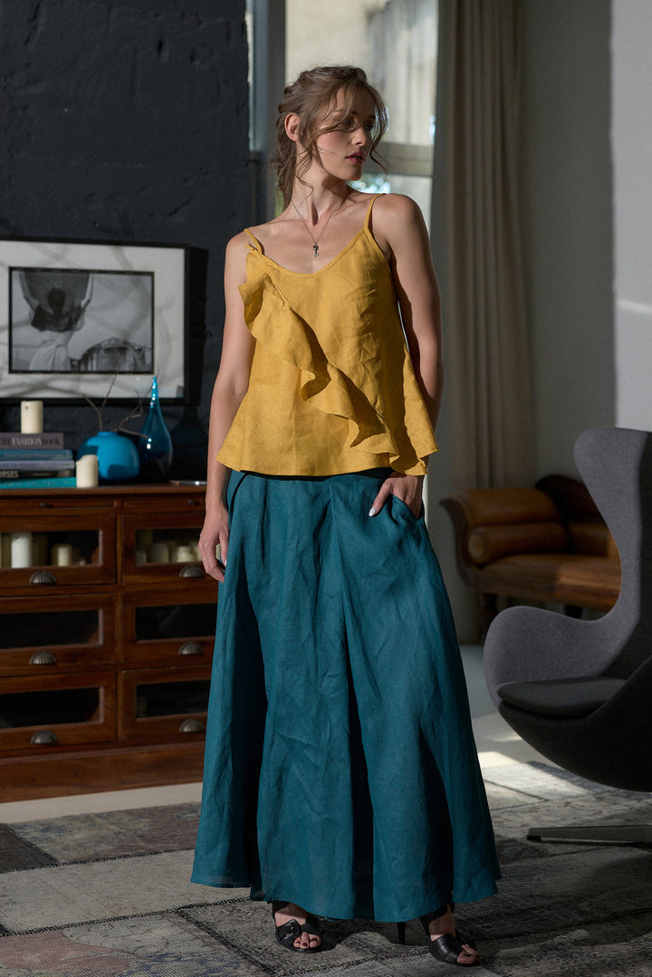 A woman stands in a modern room wearing a mustard yellow ruffled camisole top and a teal blue wide-leg skirt. The room features a dark brick wall, a wooden cabinet, and a gray chair.