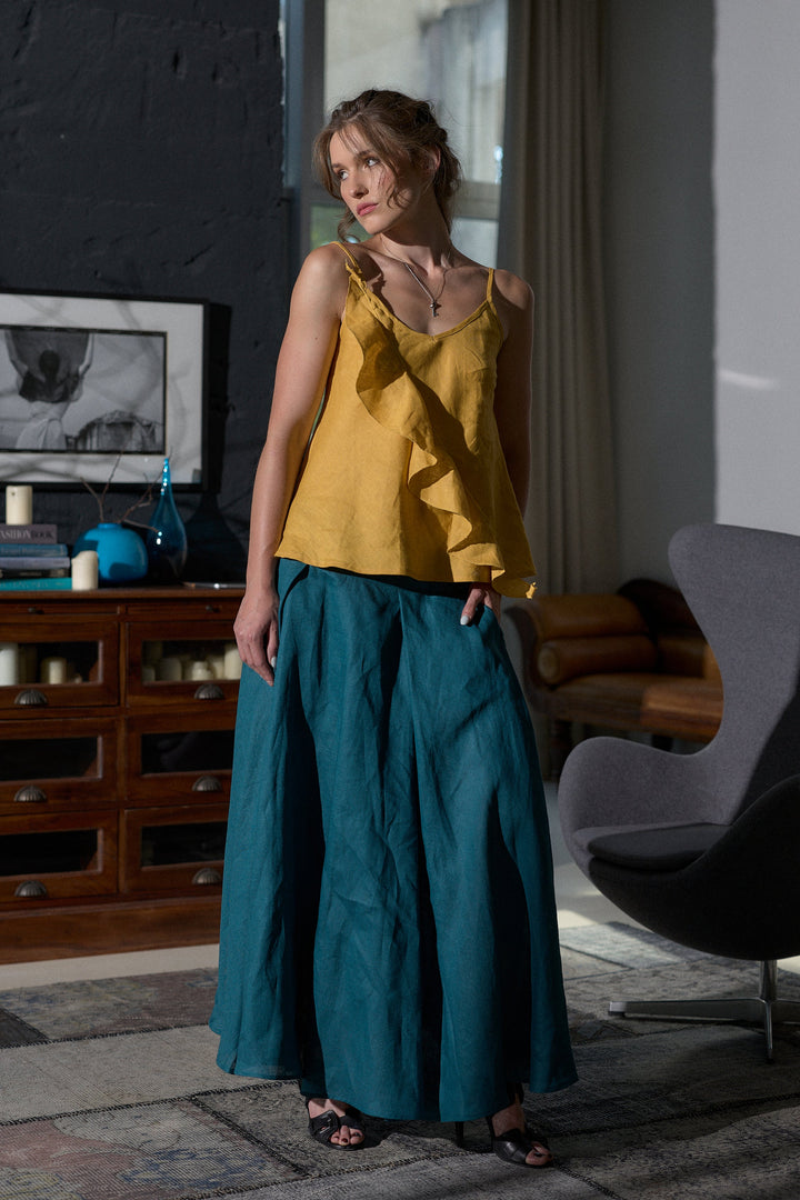 A woman modeling a mustard yellow ruffled camisole top and teal wide-leg linen pants in a modern, stylishly decorated room with dark brick walls and a wooden cabinet.