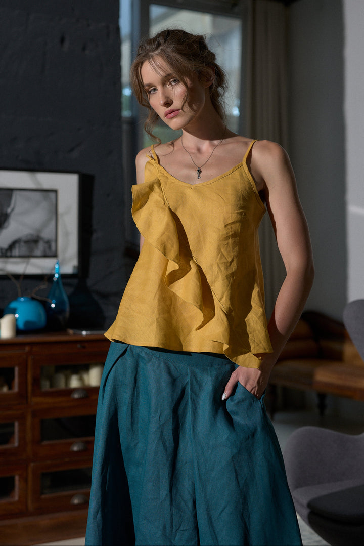 A woman models a mustard yellow ruffled camisole top paired with a teal skirt in a modern, dimly lit room with dark walls and wooden furniture.