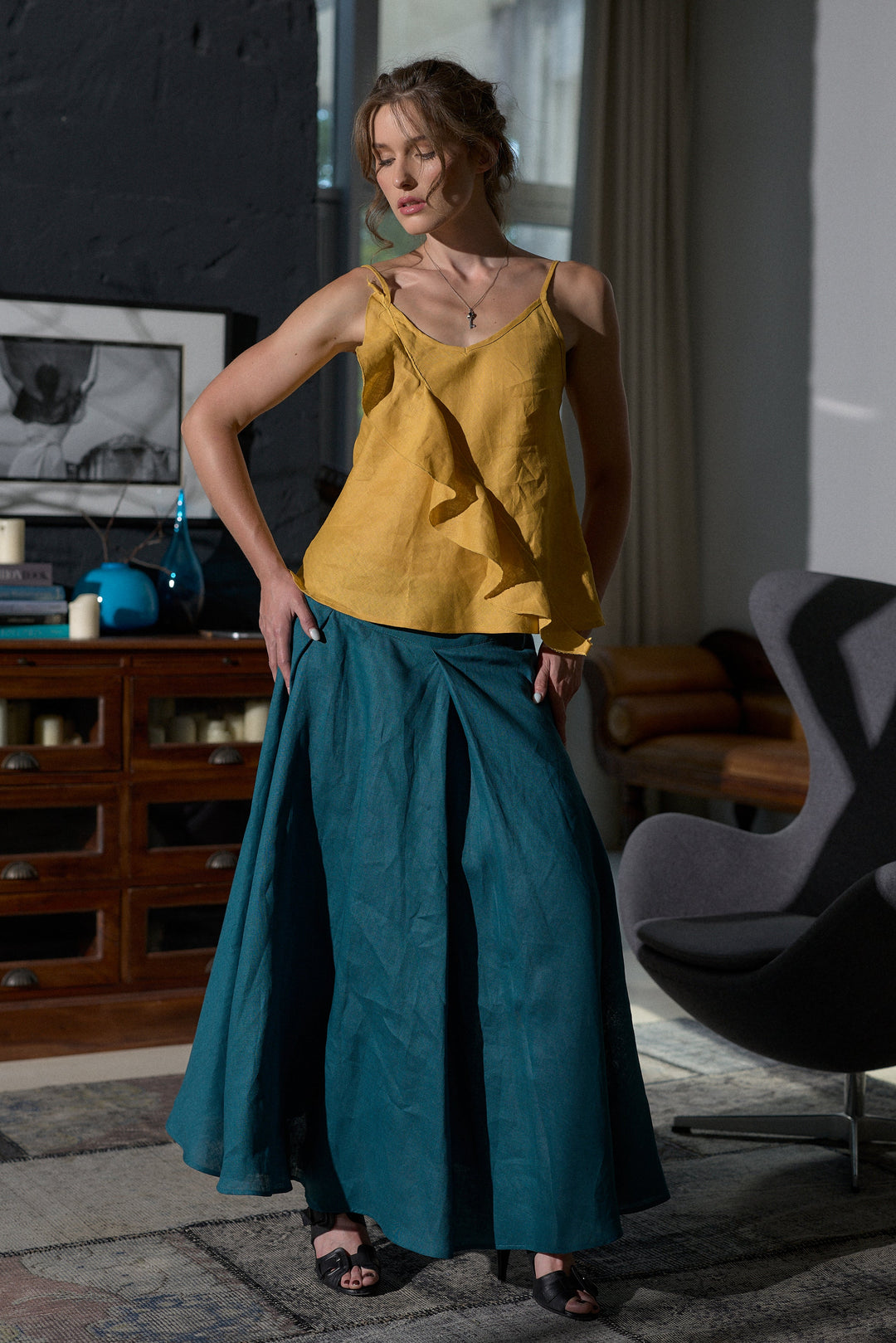 A woman modeling a two-piece outfit consisting of a mustard yellow ruffled camisole top and a teal blue wide-leg skirt, standing in a modern, stylishly decorated room with a dark gray wall, wooden cabinet, and gray chair.