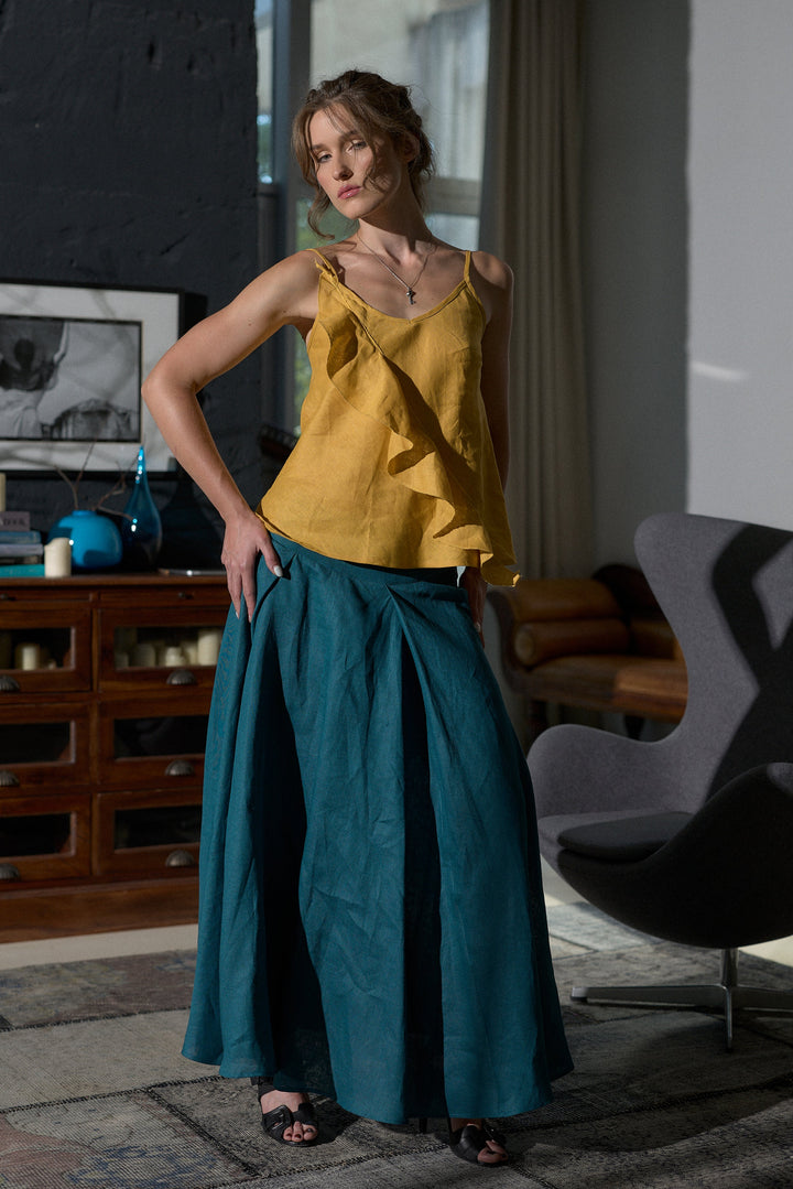 A woman models a two-piece outfit consisting of a mustard yellow ruffled sleeveless top and a teal wide-leg skirt, standing in a modern, sunlit room with a dark wood cabinet and a gray chair.