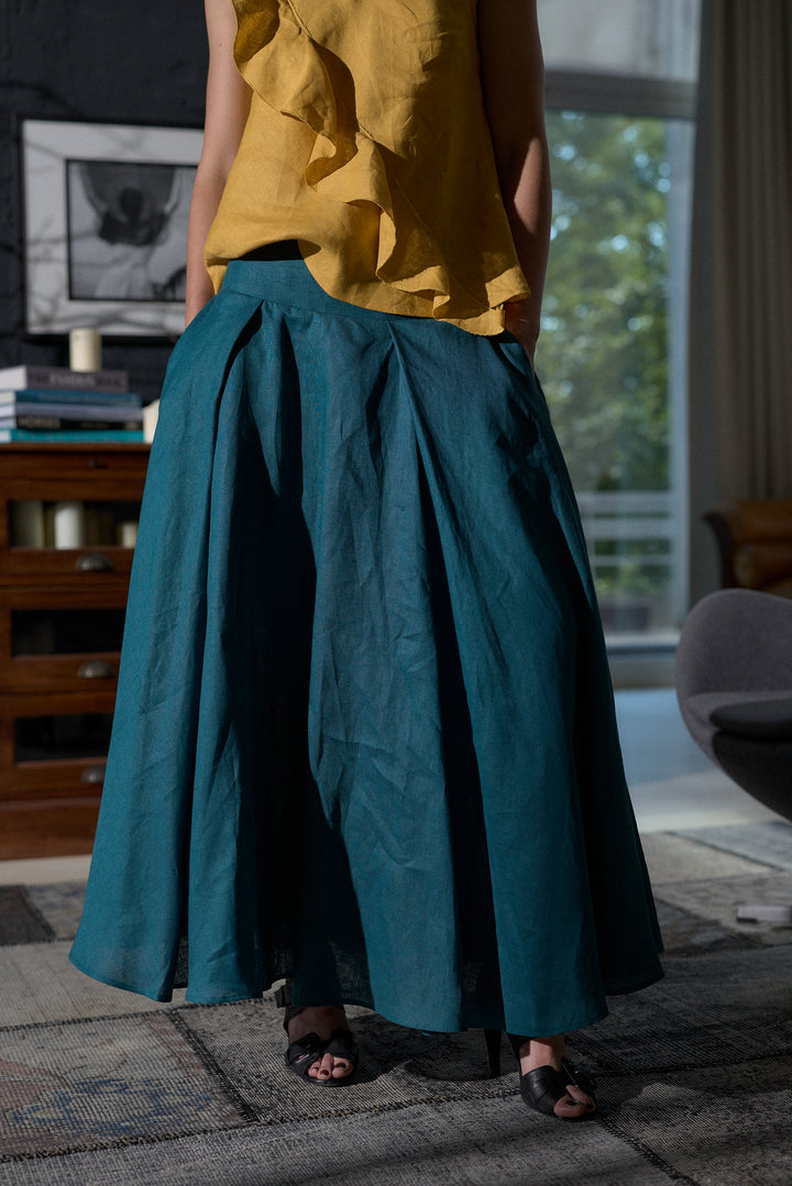A person wearing a voluminous pleated maxi skirt in deep teal blue-green, paired with a mustard yellow ruffled top and black sandals, standing in a modern living room.