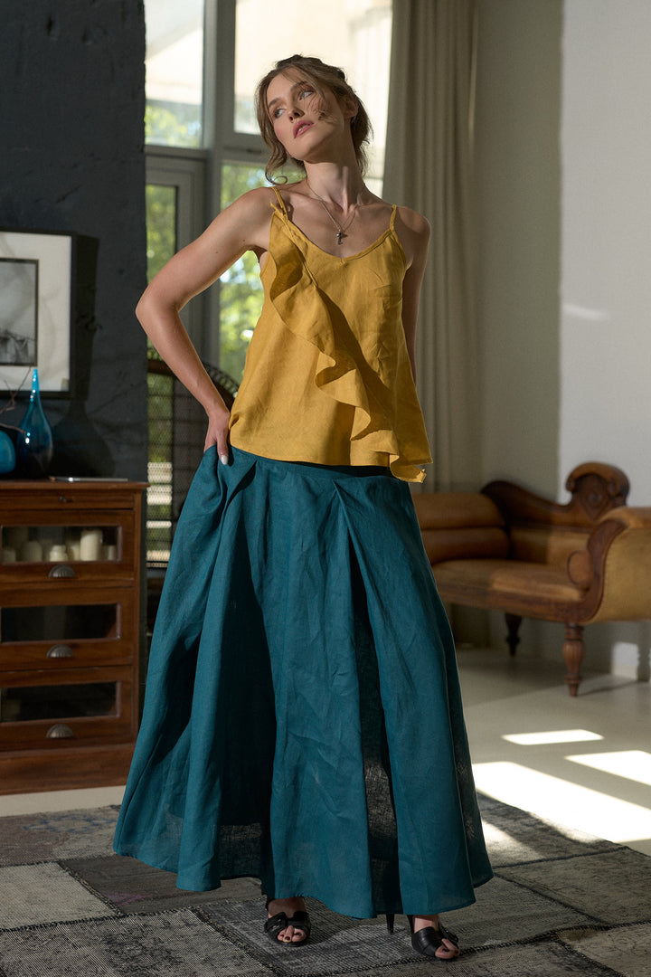 A woman modeling a mustard yellow ruffled camisole top and a teal wide-leg skirt in a sunlit room with a window, wooden furniture, and a sofa.