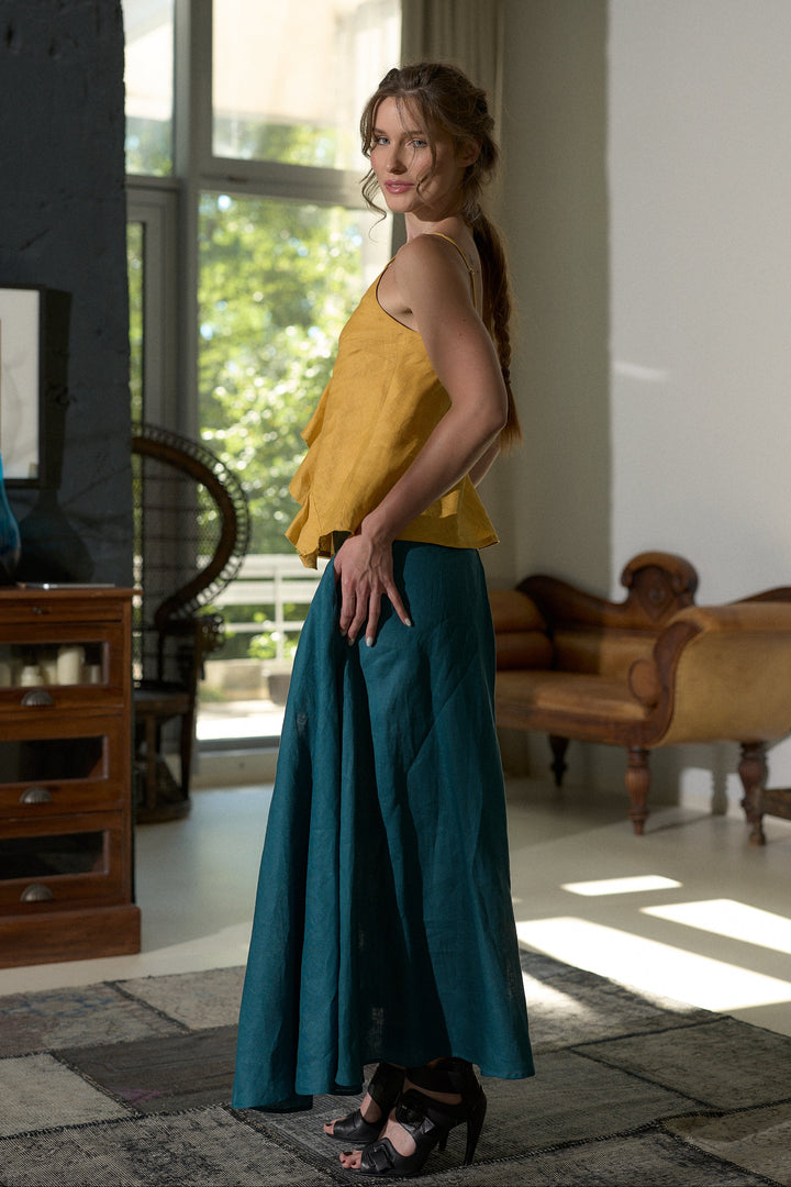 A woman stands in a sunlit room wearing a mustard yellow sleeveless top and a long teal skirt, with black heeled sandals. The room has large windows, a wooden cabinet, and a tan upholstered chair.