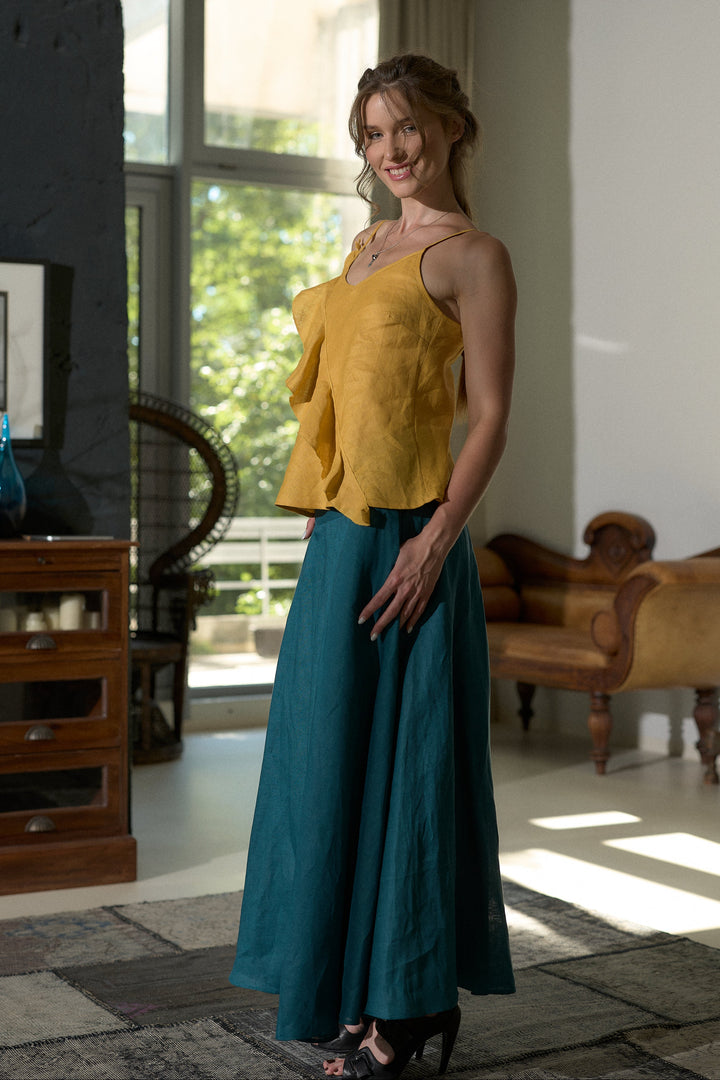 A woman modeling a two-piece outfit consisting of a mustard yellow ruffled sleeveless top and a teal blue maxi skirt, standing in a modern, sunlit room with a window and wooden furniture.