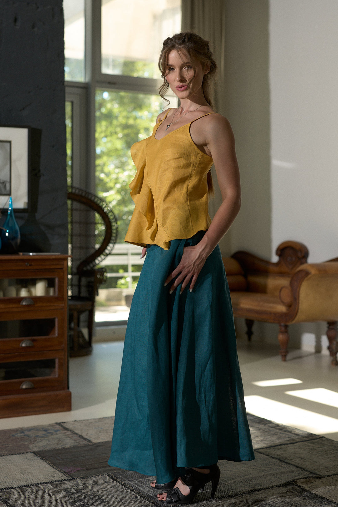 A woman models a two-piece outfit consisting of a mustard yellow ruffled sleeveless top and a teal wide-leg skirt, standing in a modern, sunlit room with wooden furniture.
