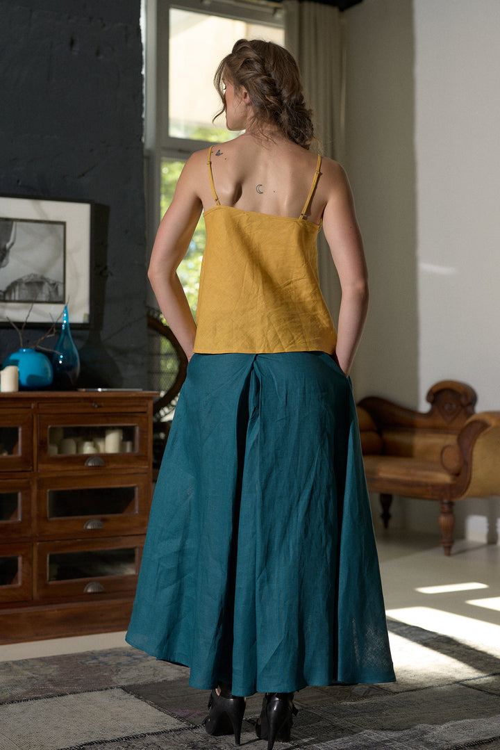 A woman stands with her back to the camera wearing a two-piece linen set consisting of a mustard yellow spaghetti-strap top and a teal blue flared skirt, in a modern room setting.