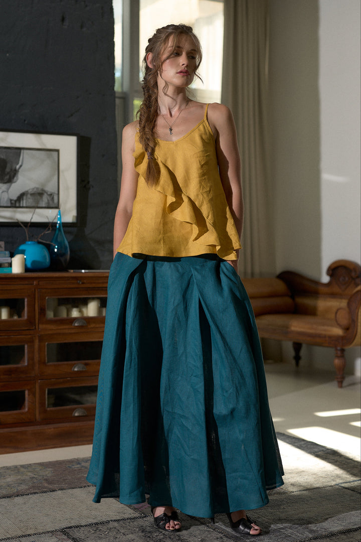 A woman stands indoors wearing a mustard yellow ruffled camisole top and a voluminous teal wide-leg skirt. The room has a dark wood cabinet, a framed print, a window with curtains, and a wooden sofa in the background.