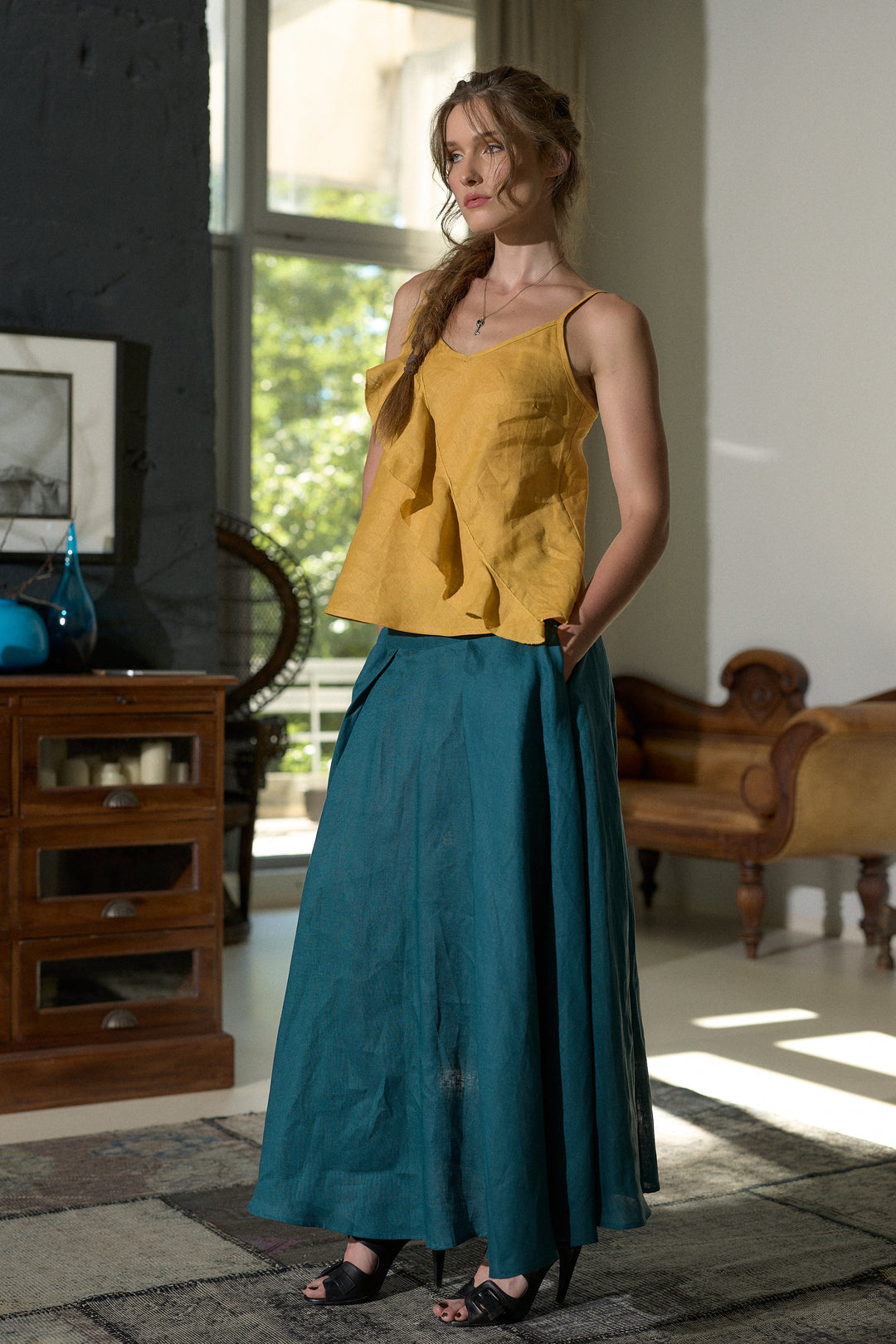 A woman stands indoors wearing a mustard yellow ruffled sleeveless top and a long teal skirt. The room has dark and light walls, a window with daylight, and wooden furniture including a cabinet and an armchair.