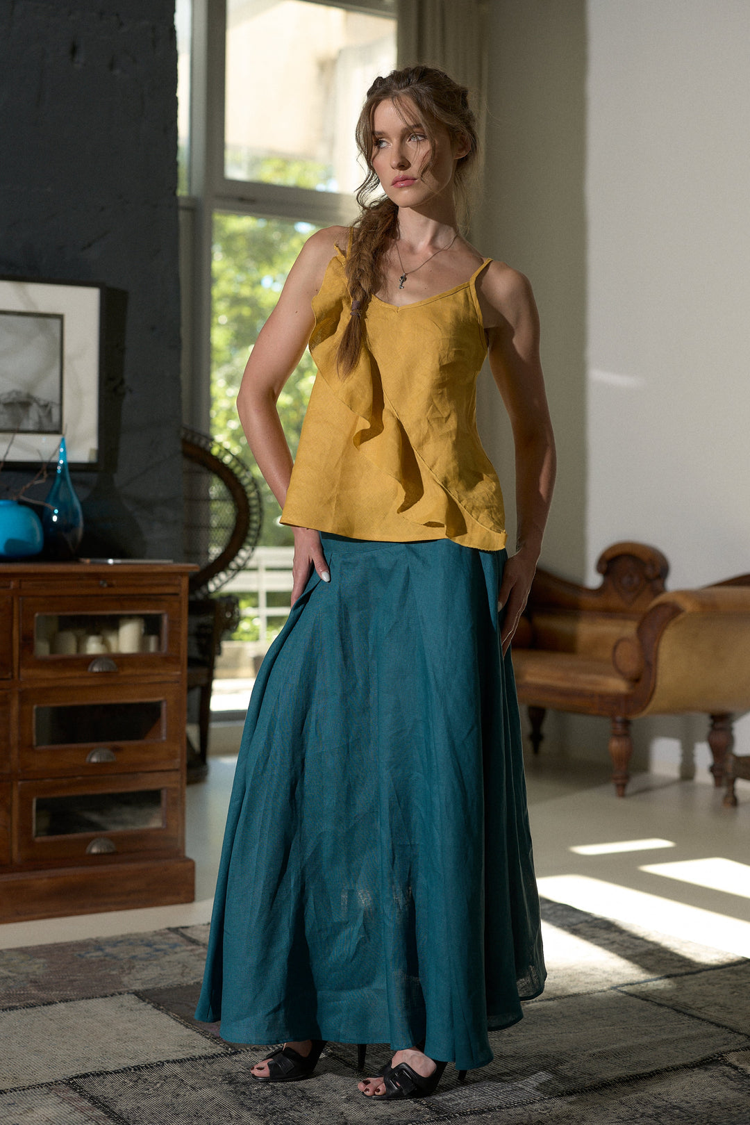 A woman stands indoors wearing a mustard yellow ruffled camisole top and a long teal skirt. The room has a dark wood cabinet, a window, and a wooden armchair, with sunlight streaming in from the side.