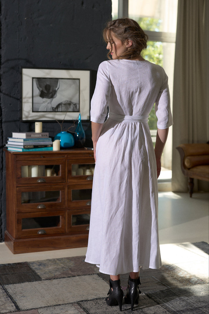 A woman wearing a light lavender linen midi dress with three-quarter sleeves and a tie waist, standing in a room with a dark wood cabinet and a window in the background.
