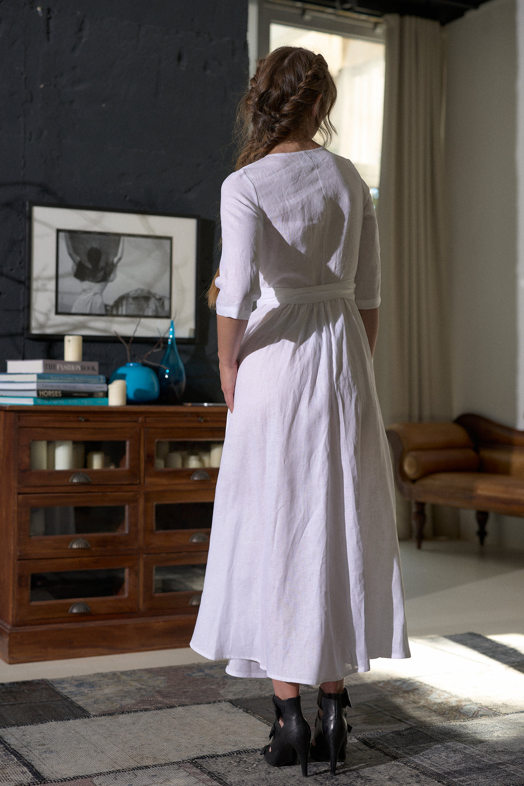 A woman viewed from behind wearing a white linen midi dress with three-quarter sleeves and a tie waist, standing in a rustic room with a dark wood cabinet and dark wall.