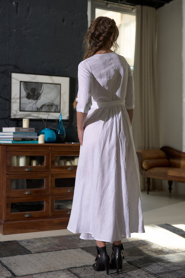 A woman viewed from behind wearing a white linen midi dress with three-quarter sleeves and a tie waist, standing in a rustic room with a dark wood cabinet and dark wall.