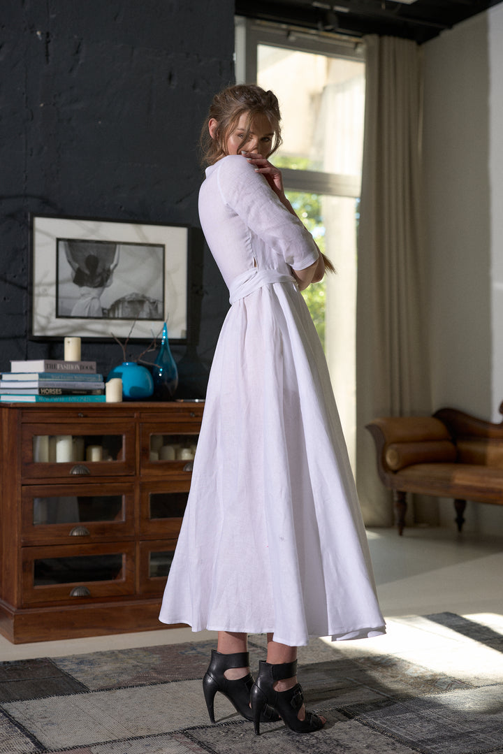 A woman wearing a white midi dress with three-quarter sleeves and a matching waist tie, standing in a modern room with a dark wood cabinet and window in the background.