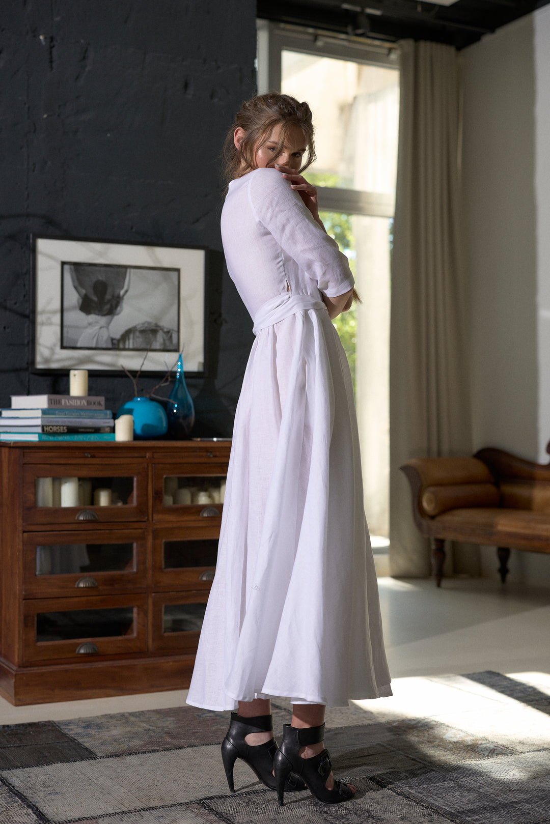 A woman wearing a long-sleeved white midi dress with a matching waist tie and black high-heeled sandals, standing in a modern room with a dark textured wall, wooden cabinet, and window.