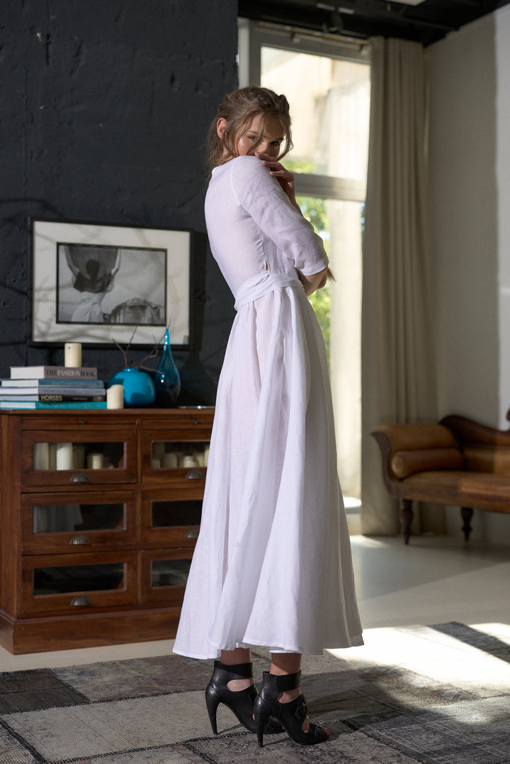 A woman wearing a long-sleeved white midi dress with a matching waist tie and black high-heeled sandals, standing in a modern room with a dark textured wall, wooden cabinet, and window.