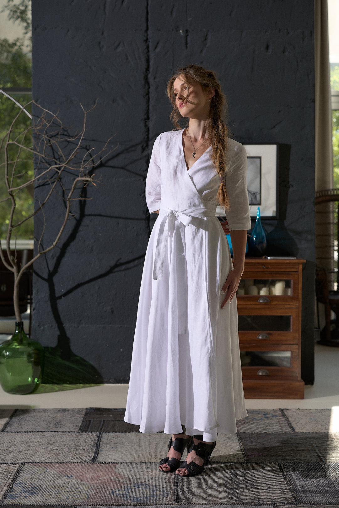 A woman wearing a white long-sleeve wrap maxi dress with a tie waist, standing in a modern interior setting with a dark textured wall and wooden furniture in the background.
