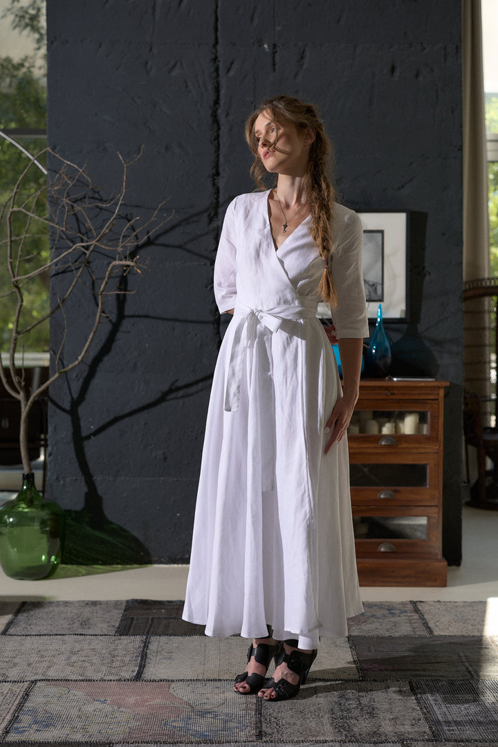 A woman wearing a white long-sleeve wrap maxi dress with a tie waist, standing in a modern interior setting with a dark textured wall and wooden furniture in the background.