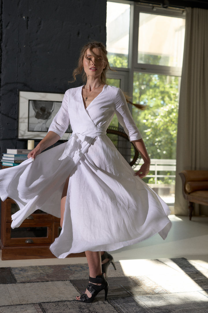 A woman wearing a white wrap-style midi dress with three-quarter sleeves and a tied waist belt, captured mid-spin in a sunlit room with large windows and dark textured walls.