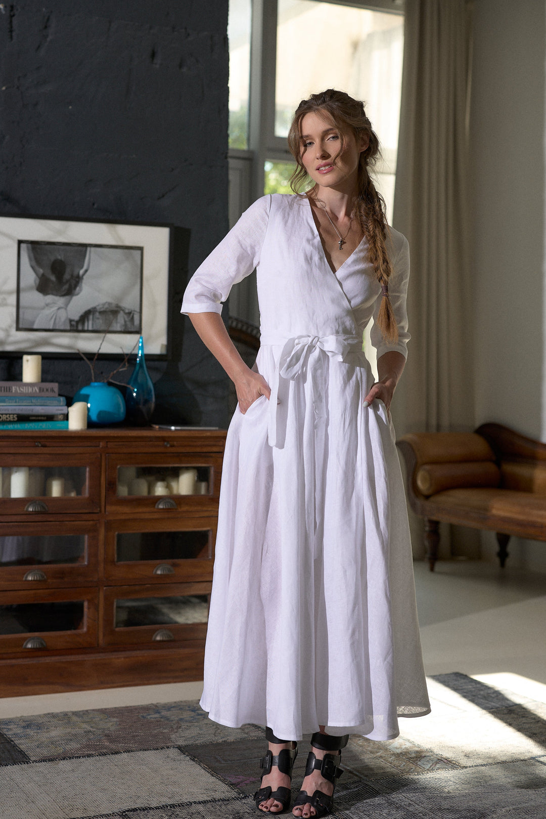A woman modeling a white midi wrap dress with three-quarter sleeves and side pockets, standing in a modern room with a dark wood cabinet and textured walls.