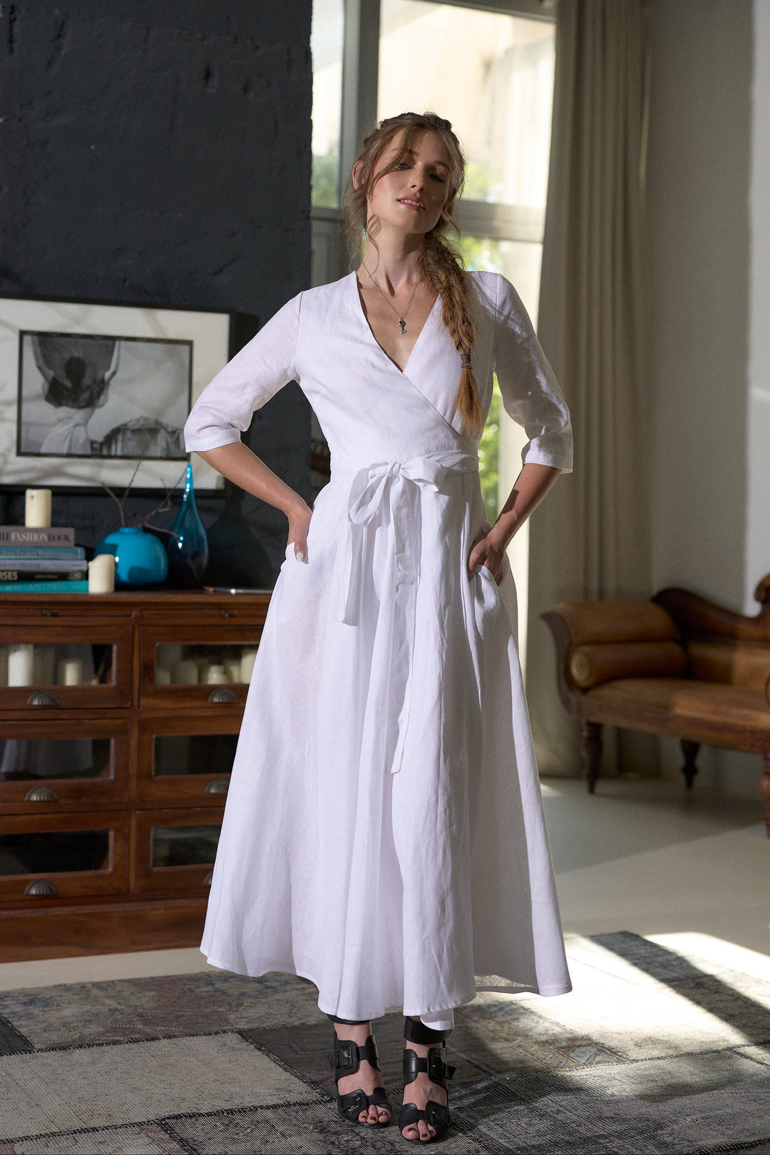 A woman modeling a white midi wrap dress with three-quarter sleeves and a tie waist, standing in a modern room with a dark wood cabinet and window.