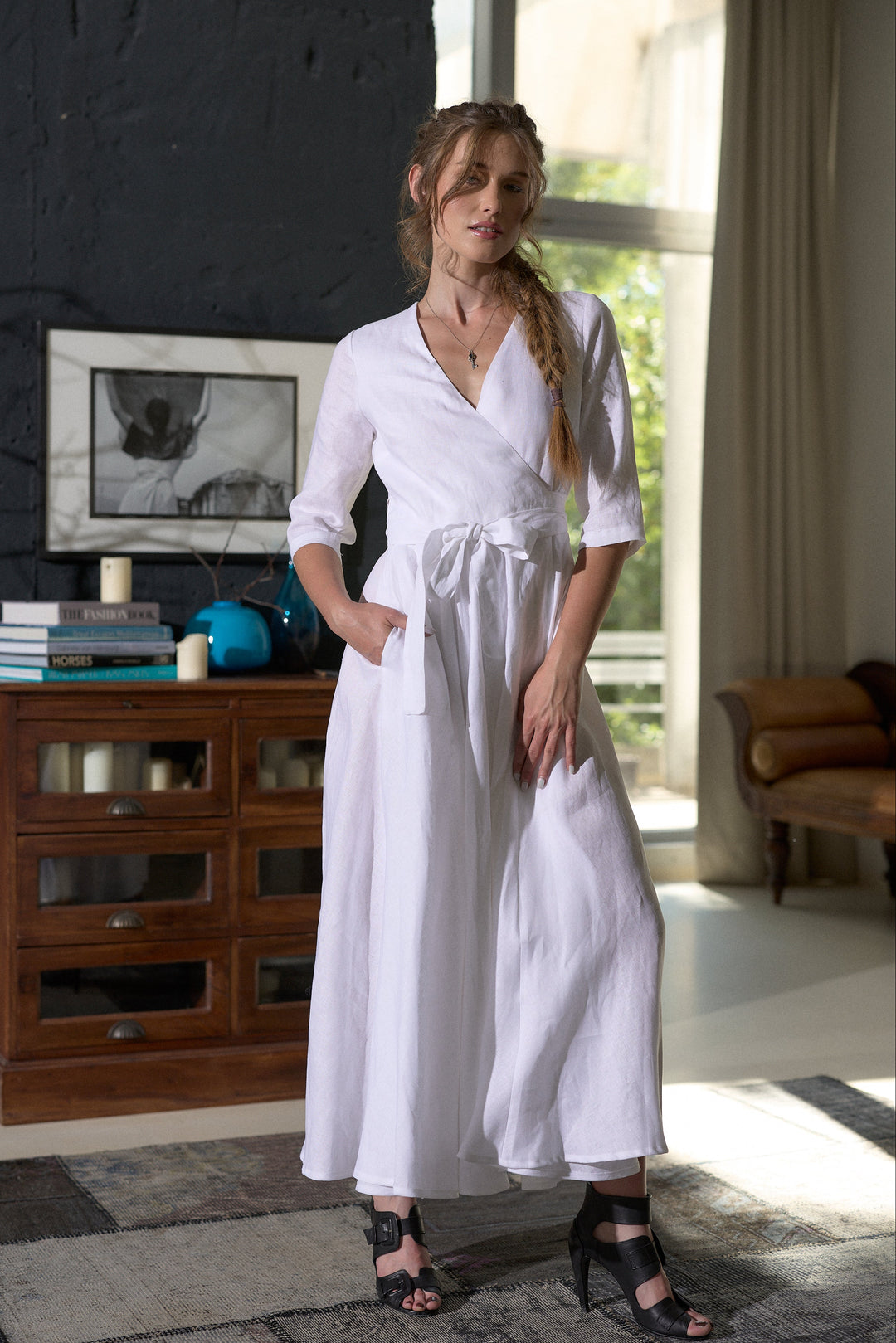 A woman models a white linen wrap dress with three-quarter sleeves and a tie waist, standing in a modern room with a dark brick wall and a wooden cabinet.