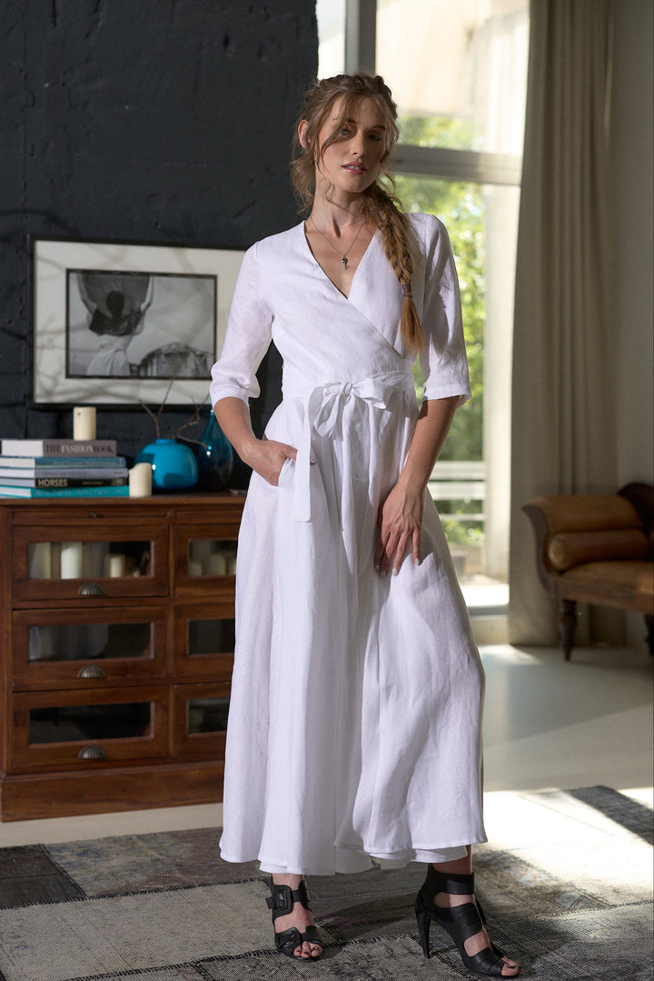 A woman models a white linen wrap dress with three-quarter sleeves and a tie waist, standing in a modern room with a dark brick wall and a wooden cabinet.