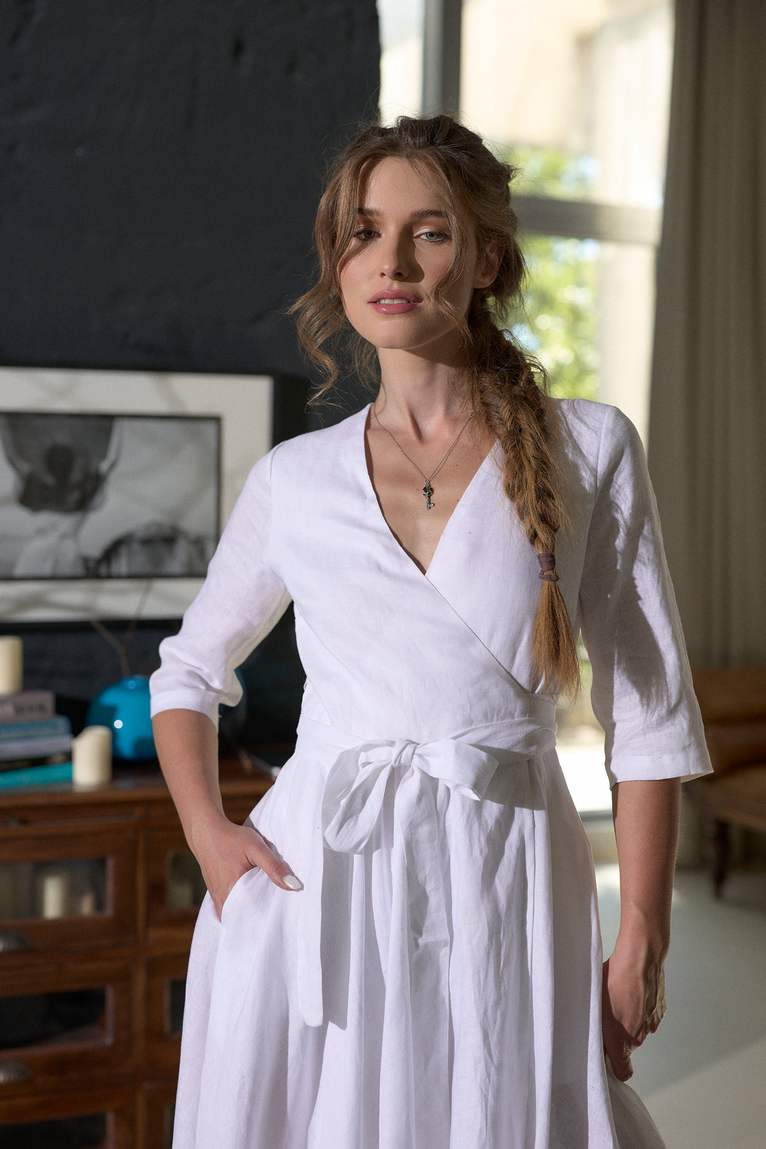 A woman wearing a white linen wrap dress with three-quarter sleeves and a self-tie waist, standing in a softly lit room with a dark wall and wooden furniture in the background.