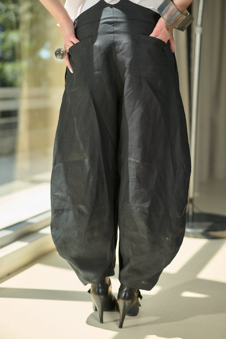 A person shown from the back wearing loose, voluminous black harem-style pants with side pockets, paired with black pointed heels in a bright indoor setting.