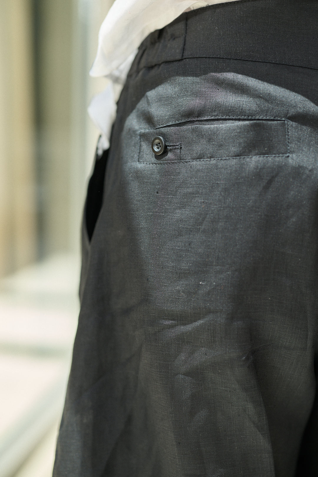 Close-up of dark gray trousers showing the back pocket with a button closure, worn with a white shirt.